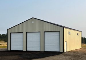 A beige metal building with three large white garage doors and a small window on the side, situated on a dirt lot.