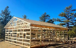 A wooden frame structure of a building under construction stands on a cleared area, surrounded by trees and a clear blue sky.