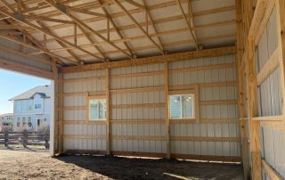Interior view of an unfinished wooden and metal barn or shed with exposed beams, dirt floor, two windows, and part of a house visible outside.