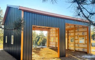 A partially constructed metal building with open garage-style entrances, exposed framing, and visible insulation, set on a dirt lot surrounded by trees.