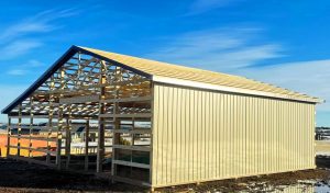 A partially constructed building with exposed wooden framing and a metal exterior stands on a dirt lot under a blue sky.