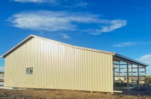 A beige metal building with a sloped roof stands on dirt ground under a blue sky; one side is partially open with visible support beams.