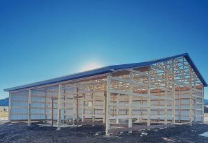 A partially constructed metal and wood-framed building stands on a dirt lot under a clear blue sky, with sunlight behind the structure.