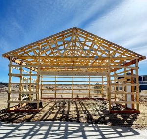 A wooden house frame stands on a concrete foundation in a sandy, open area under a blue sky with wispy clouds.