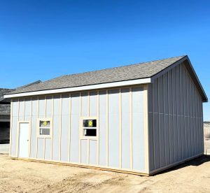 A light gray shed with a sloped roof, two small windows, and a door, standing on a dirt lot under a clear blue sky.