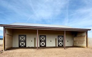 A tan metal barn with a gray roof features three stall doors and wooden side walls, set on a dry, dirt ground under a blue sky with light clouds.