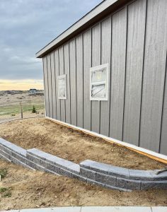 A gray shed with two small windows is next to a partially finished retaining wall and a patch of bare soil. The surrounding area is open and grassy with some distant houses.