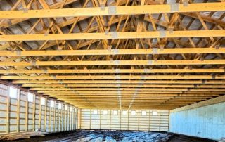 Interior view of a large unfinished building with exposed wooden roof trusses, open walls, and a dirt floor.