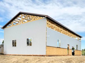 A partially constructed building with exposed roof trusses, unfinished walls, and several installed windows, set on a dirt lot under a partly cloudy sky.