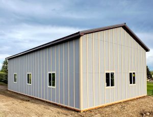A large, light gray metal building with a dark roof, vertical siding, and several rectangular windows, situated on bare ground under a partly cloudy sky.