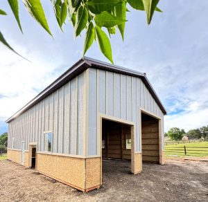 A large, unfinished metal and wood barn with two wide door openings and one window stands on a dirt lot under a partly cloudy sky.