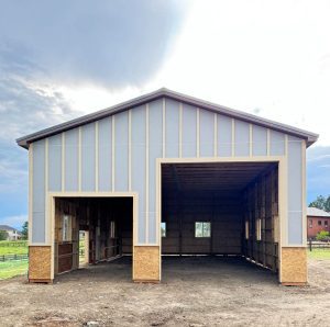 A large, unfinished barn with open entryways, visible framing, and no doors, set on a dirt ground with some grass and buildings in the background.