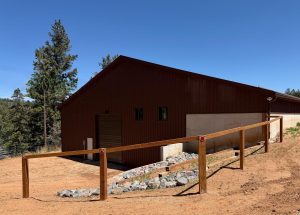 A brown wooden building with a sloped roof stands on a dirt lot, bordered by a wooden fence and a rock-lined drainage path, with trees and blue sky in the background.