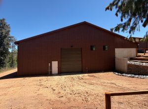 A large brown metal building with a closed garage door, two small windows, and a dirt lot in front on a sunny day.
