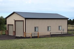 A beige metal building with a large garage door, several windows, and a fenced yard stands on a grassy property.