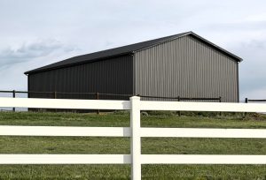 A large gray metal barn sits behind a white fence on a grassy field under a cloudy sky.