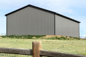 A large, gray metal building with a pitched roof stands on a grassy slope behind a wooden fence under a partly cloudy sky.