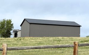 A large gray metal barn with a black roof stands on a grassy hill, partially fenced in by a wooden and wire fence under a cloudy sky.