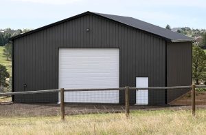 A large dark gray metal building with a white garage door and a white entry door, situated behind a fenced grassy area.