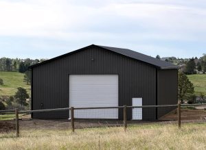 A large, dark gray metal building with a white garage door and adjacent entry door, set on a grassy lot with a wooden fence in the foreground.