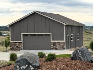 Gray detached garage with a gable roof, stone accents, and two windows, set on a concrete driveway with landscaped rocks and grass in the foreground.