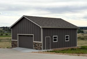 A brown detached garage with a single overhead door, two small windows, and stone accents at the base, situated on a concrete driveway in a rural area.