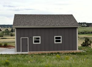 A small gray building with a gable roof, two rectangular windows, and a door, situated in a grassy open area with fields and trees in the background.