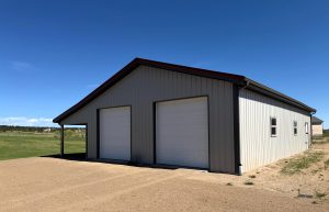 A large metal building with two white garage doors stands on a dirt driveway in a rural area under a clear blue sky.