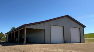 A large metal building with two white garage doors and an open side shelter stands on a dirt lot under a clear blue sky.
