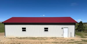 A rectangular metal building with a red roof, two windows, and a white door, situated on a grassy landscape under a clear blue sky.