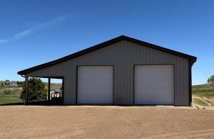 A metal building with two white garage doors stands on a gravel lot under a clear blue sky, with grassy fields and scattered trees in the background.
