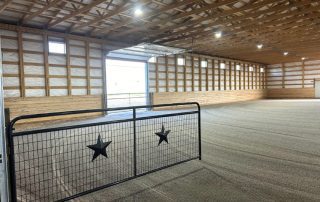 Interior of a spacious indoor riding arena with riding arena dust control, featuring a metal gate with star cutouts, gravel floor, and wooden support beams beneath a high ceiling.