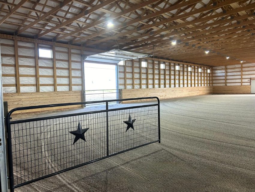 Interior of a spacious indoor riding arena with riding arena dust control, featuring a metal gate with star cutouts, gravel floor, and wooden support beams beneath a high ceiling.