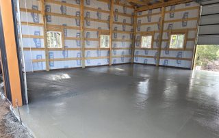 A freshly poured concrete floor inside a partially constructed building with unfinished insulation and exposed wooden framing.