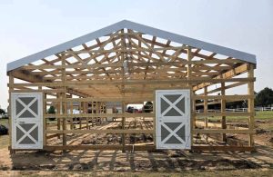 A partially constructed wooden barn frame with two double doors, set on a dirt ground in an open outdoor area.
