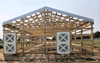 A partially constructed wooden barn frame with two double doors, set on a dirt ground in an open outdoor area.