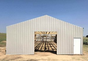 A metal building frame with an open garage door and a closed white entry door, showing interior wooden beams, stands on a dirt lot under a clear sky.