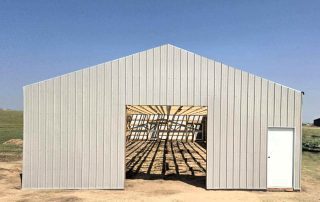 A metal building frame with an open garage door and a closed white entry door, showing interior wooden beams, stands on a dirt lot under a clear sky.