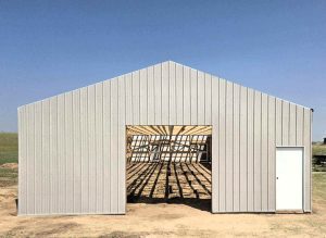 A metal building frame with a large open entrance and a closed door stands on dirt ground, casting shadows inside under a clear blue sky.