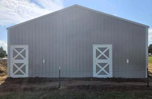 A large, light gray barn with a peaked roof and two white, X-braced doors, situated on a dirt and grassy area under a partly cloudy sky.