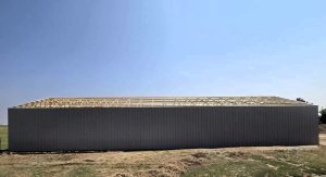 A large rectangular building with metal siding and an unfinished roof frame stands on bare ground under a clear blue sky.