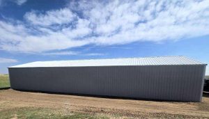A large, rectangular metal building with a sloped roof stands on a dirt lot under a partly cloudy sky.