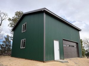 A green metal building with a pitched roof, two windows, a white door, and a brown garage door, situated on sandy ground with trees in the background.