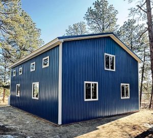 A blue, two-story metal-sided building with multiple white-trimmed windows, situated in a wooded area with tall pine trees.