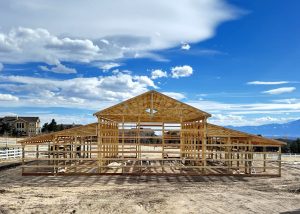 A wooden barn frame under construction stands on a dirt lot, with houses, fencing, and mountains visible in the background under a partly cloudy sky.