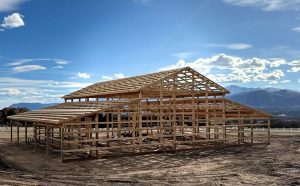 A wooden frame of a large building under construction stands on a dirt lot, with mountains and blue sky in the background.