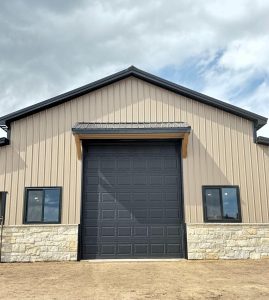 A beige metal building with a large black garage door and two windows, set on a stone foundation under a cloudy sky.