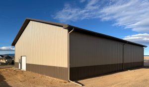 Large beige and brown metal-sided warehouse with a single white door, situated on bare dirt under a partly cloudy sky.