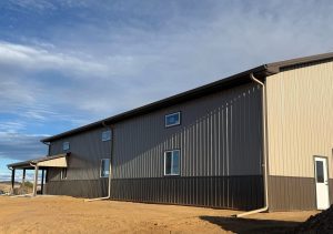 A large metal building with multiple windows and doors sits on a dirt lot under a partly cloudy sky.