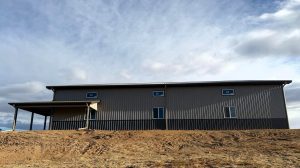 A large gray metal building with a covered porch stands on bare, uneven ground under a partly cloudy sky.
